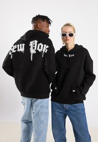 Felpe nere con cappuccio che presentano la scritta "New York" in bianco, con un design di carattere gocciolante. Abbinamento di jeans azzurri e blu scuro, che mostrano un fit rilassato.