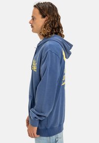 Modrá bavlnená mikina s kapucňou, predné vrecko, žltý grafický nápis na hrudi a šnúrka na kapucni. Hladká textúra, voľný strih.