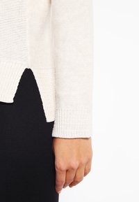 Nahaufnahme einer Person, deren Hand neben dem Bund und dem seitlichen Schlitz eines cremefarbenen Strickpullis über schwarzen Hosen liegt.