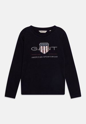 Granatowa koszulka z długim rękawem wykonana z bawełny. Posiada logo w kształcie tarczy z czerwono-białymi paskami oraz szary napis "GANT" nad napisem "AMERICAN SPORTSWEAR."
