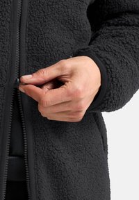 Schwarze Fleecejacke mit einer strukturierten Oberfläche, ausgestattet mit einem Reißverschluss vorne und enganliegenden Bündchen. Es ist zu sehen, wie eine Hand den Reißverschluss zieht.