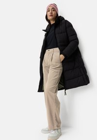 Black puffer coat with a hood, paired with beige corduroy pants and light gray sneakers. The model wears a pink headband.