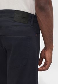 Κοντινό πλάνο ενός ατόμου που φορά σκούρο μπλε παντελόνι με μαύρη ετικέτα Jack & Jones στη μέση και ένα λευκό πουκάμισο μέσα από το παντελόνι.