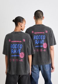 Pelēka oversized T-krekls ar drosmīgu balto, rozā un zilo grafisko tekstu aizmugurē, tostarp "Rodeo Rave" un smaidoša sejiņa.