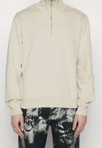 Sudadera cremosa con cremallera y cuello alto, textura suave y un logotipo minimalista en el lado izquierdo del pecho, combinada con unos vaqueros negros estampados.
