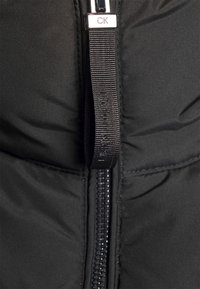 Fermeture éclair noire sur tissu noir avec une tirette noire portant le logo embossé Calvin Klein et une pièce métallique gravée CK.