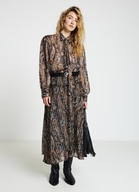 Robe longue à motifs dans des tons noirs, marron et bleu avec un col noué, taille cintrée et une silhouette fluide ; associée à des bottes noires et un sac.