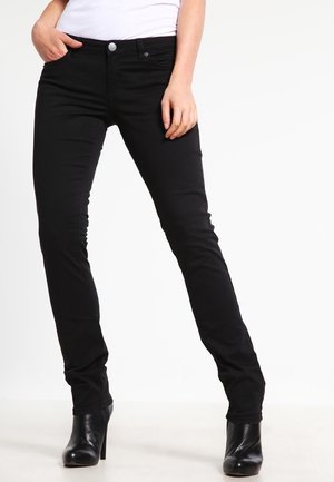 Femme portant un jean skinny noir et des bottines à talons noires, une main dans la poche et l'autre reposant sur la cuisse.