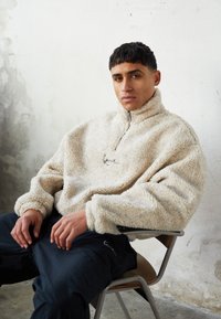 Beige fleece genser med høy krage, glidelåslukking og teksturert overflate. Modellen har på seg mørke bukser og sitter på en stol mot en nøytral bakgrunn.