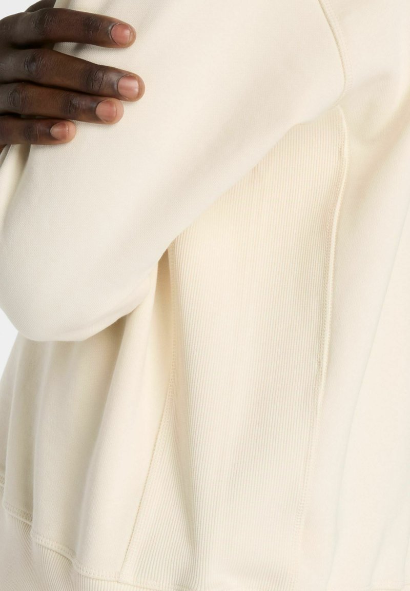 Hellbeiges Sweatshirt aus weichem Stoff, mit einem Rippdetail auf einer Seite und einem lockeren Sitz mit sichtbaren Nähten. Hände sind teilweise sichtbar.