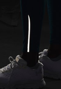 Mörka sportbyxor med reflekterande söm på sidan; ihop med vita sportskor som visar nätstruktur och logotypdetalj.