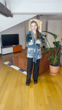 Modrý pletený kardigan s více vzory a béžovými akcenty, zavázaný v pase, doplněný černými kalhotami a tmavými botami, v moderním vnitřním prostředí.