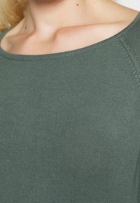 Dunkelgrüner, gerippter Stoff mit einem U-Boot-Ausschnitt, der eine weiche Textur und glatte Nähte aufweist. Nahaufnahme, die das Material hervorhebt.