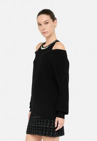 Žena u crnom oversized puloveru s otvorenim ramenima preko bezrukavog crnog topa sa srebrnim detaljima i crnom mini suknjom s uzorkom.