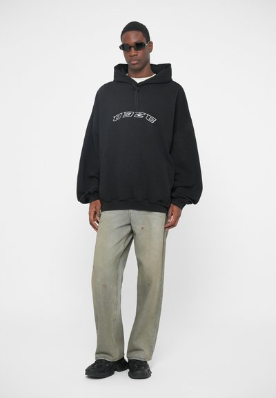 Μαύρη oversized φούτερ με μπροστινή εκτύπωση, ανοιχτό πράσινο τζιν με φθορές και μαύρα αθλητικά παπούτσια. Το μοντέλο φοράει σκούρα γυαλιά ηλίου και στέκεται σε έναν απλό φόντο.