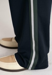 Marineblauwe broek met witte en donkergroene zijstrepen, gedragen met crèmekleurige geweven leren schoenen op een lichte vloer.