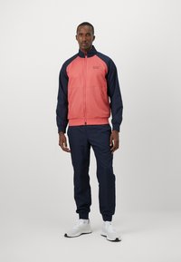 Ensemble de survêtement composé d'une veste zippée corail avec des manches bleu marine et un pantalon bleu marine assorti. Fabriqué en tissu lisse, avec des poignets et un col côtelés.