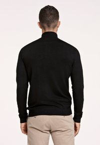 Maglione nero a collo alto in maglia con maniche lunghe, polsini e orlo a coste, indossato con pantaloni beige chiaro. La texture sembra liscia e aderente.