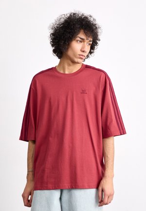 Rotes übergroßes T-Shirt aus Baumwolle mit kurzen Ärmeln, das ein kleines Branding-Logo auf der Brust und drei dunkle Streifen auf jeder Schulter aufweist.
