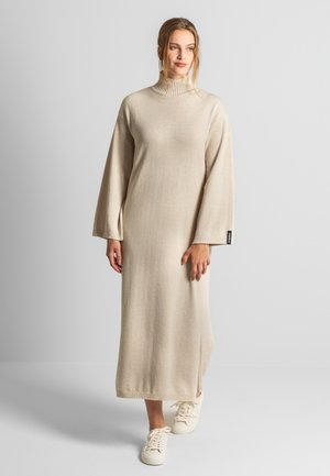 Beiges langes Strickkleid mit ribbelt rotem hohen Ausschnitt und weiten Ärmeln, mit Seitenschlitzen, kombiniert mit weißen Sneakers für den Kontrast.