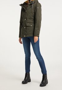 Person som bär en olivgrön huva med stora knappar, blå skinny jeans och svarta snörstövlar, stående mot en enfärgad bakgrund.