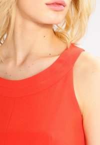 Červený bezrukávový top s hladkou texturou a kulatým výstřihem. Materiál vypadá lehce, s čistým a jednoduchým designem.