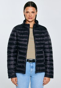 Vrouw draagt een zwarte gewatteerde jas met ritssluiting over een bruin-wit gestreept shirt, lichte blauwe spijkerbroek en een zwarte riem.