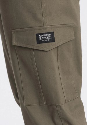 Oliivinvihreät cargo-housut, joissa on myyntipussitasku mustalla etiketillä, jossa lukee "AINA RAJASI ON MIELI", siistillä tikillä.