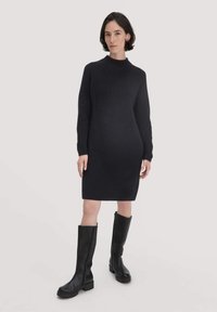 Schwarzes gestricktes Kleid mit hohem Kragen, langen Ärmeln und figurbetontem Schnitt. Kombiniert mit knielangen schwarzen Lederstiefeln mit derben Sohlen.