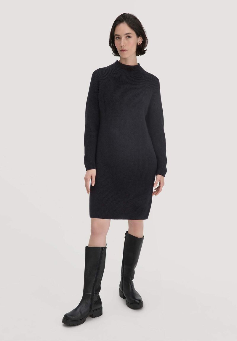 Schwarzes gestricktes Kleid mit hohem Kragen, langen Ärmeln und figurbetontem Schnitt. Kombiniert mit knielangen schwarzen Lederstiefeln mit derben Sohlen.