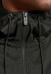 Czarna nylonowa kurtka z wysokim kołnierzem, kontrastowym zapięciem na zamek, regulowanymi sznurkami i teksturowanym wykończeniem. Logo Nike na zamku.