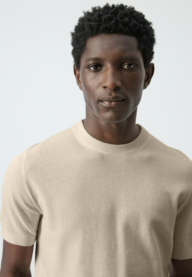 Jeune homme noir aux cheveux courts et bouclés portant une chemise en maille beige à manches courtes, debout devant un fond clair uni.