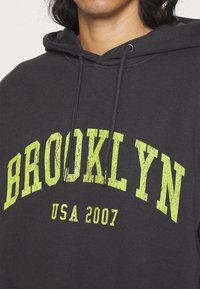 Sudadera negra con capucha y cordón, con texto grande en amarillo verdoso que dice "BROOKLYN" y "USA 2007" en un estilo de fuente agrietada.