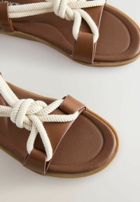 Sandales marron avec semelle rembourrée, dotées de larges sangles en corde blanche nouées sur le dessus et d’accents en cuir près des orteils et des talons.