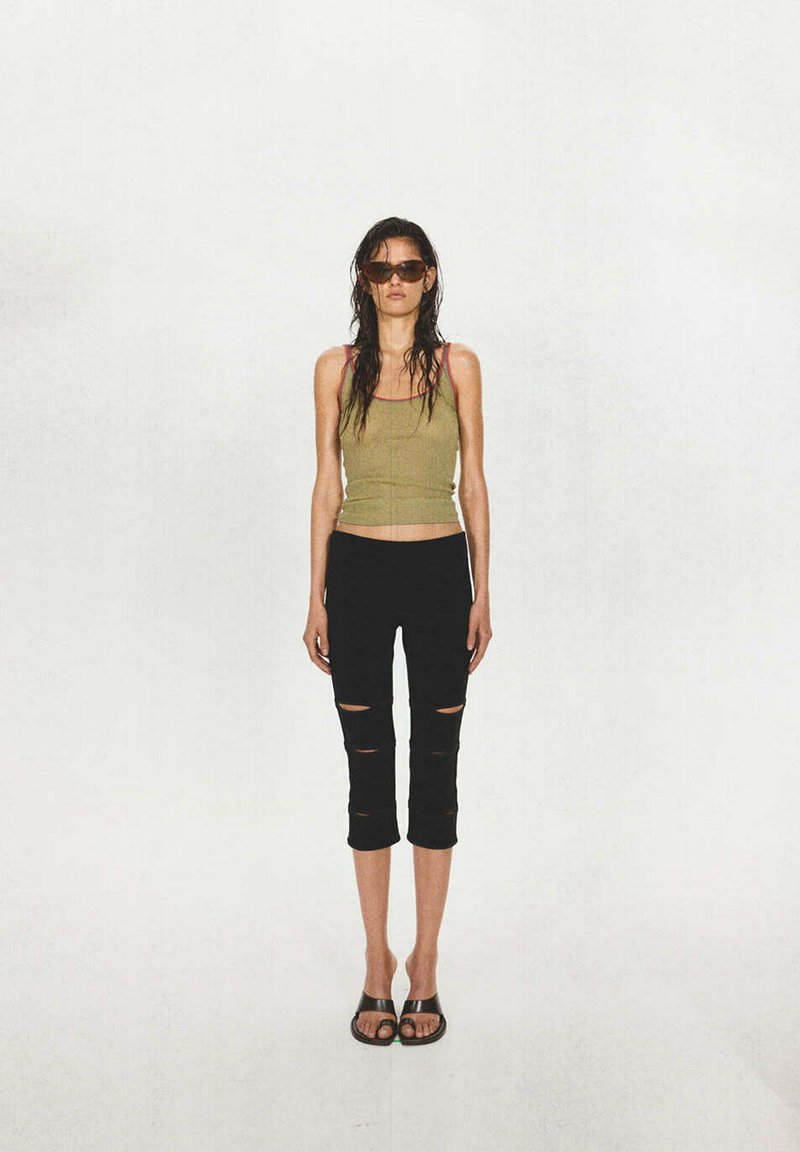 Femme aux longs cheveux mouillés, portant des lunettes de soleil, un débardeur olive, un pantalon noir déchiré et court, et des sandales noires, debout sur fond blanc.