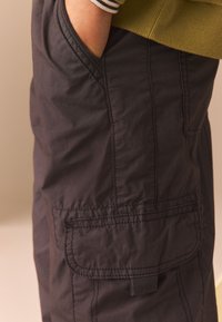 Pantaloni cargo neri realizzati in materiale leggero, con dettagli cuciti, tasche laterali e una vestibilità rilassata. Superficie testurizzata con finitura opaca.