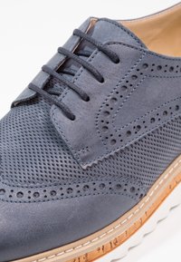 Scarpe da uomo in pelle perforata blu con chiusura con lacci, caratterizzate da una superficie testurizzata, dettagli brogue e una suola in gomma marrone chiaro.