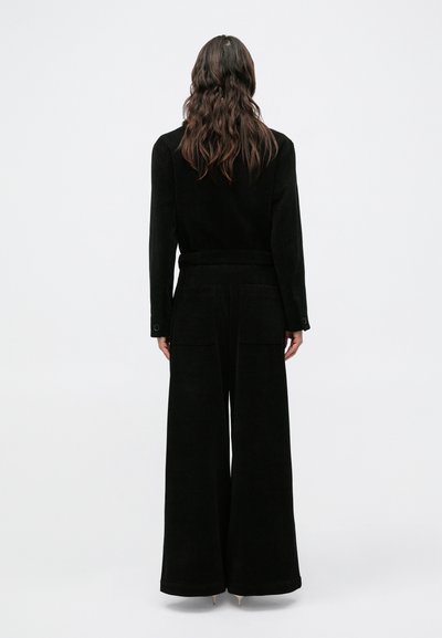 Fekete kordbársony szett széles szárú nadrággal, övvel a deréknál és két hátsó zsebbel. Látható a sima textúra és a szabott illeszkedés.