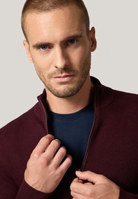 Mężczyzna z krótkimi włosami i jasną brodą zapina teksturowaną bordową kurtkę na granatową koszulę na jednolitym jasnym tle.