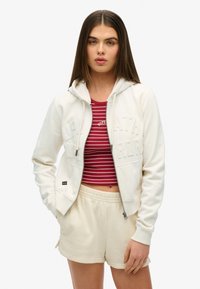Witte zip-up hoodie met textuurlettering, gecombineerd met een rood en wit gestreept crop top en crèmekleurige shorts. Glad materiaal, casual pasvorm.