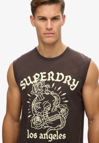 Camiseta sin mangas marrón con un estampado gráfico de una calavera entrelazada con rosas y serpientes, con "SUPERDRY los ángeles" en fuente color crema.