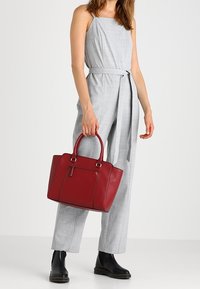 Czerwona skórzana torebka z zaokrąglonymi uchwytami, o solidnej konstrukcji, z przednią kieszenią. Modelka nosi szary kombinezon i czarne botki za kostkę.