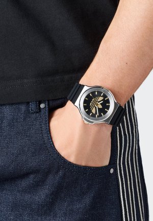Reloj con correa de goma negra, caja redonda de plata, dial negro y acentos dorados. Muñeca visible con bolsillo de tela de mezclilla y camiseta casual.
