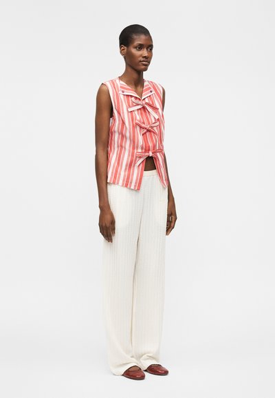 Γυναίκα με αμάνικο κόκκινο-άσπρο ριγέ τοπ με φιόγκους στο μπροστινό μέρος, φαρδιά λευκά υφασμάτινα παντελόνια και κόκκινα φλατ παπούτσια, στέκεται μπροστά σε λευκό φόντο.