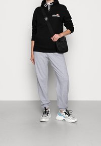 Sweat à capuche noir avec poche avant, pantalon de survêtement gris, et baskets blanches épaisses avec des accents bleus ; le modèle tient un petit sac noir.