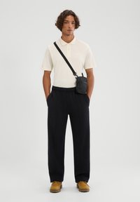 Cremefarbenes Polohemd mit kurzen Ärmeln, schwarze locker sitzende Hose, gelbe Lederstiefel und eine schwarze Umhängetasche mit strukturierter Oberfläche.