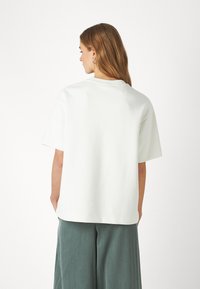 Felpa bianca a maniche corte con vestibilità comoda. Presenta un collo a giro e una texture liscia. Indossata con pantaloni larghi verdi.