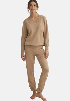 Beige gebreid loungepak met een V-hals, ribgebreide manchetten en zoom op de trui, gecombineerd met bijpassende taps toelopende broeken. Zachte textuur.