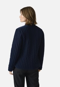 Marinblå kviltad jacka med vertikala sömmar, med rundad krage och långa ärmar. Tyget verkar mjukt och texturerat.