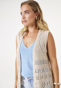 Beige gestrickte Weste mit offener Masche und gemusterten Details, über einem hellblauen Top getragen, das einen tiefen V-Ausschnitt und eine lässige Passform hat.